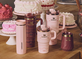 Stanley 1913 pink and burgundy tumblers and mugs with cherry designs arranged among pink and white frosted cakes.