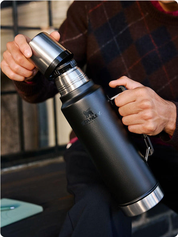 Stanley 1913 matte black insulated thermos being opened by a person wearing a plaid sweater outdoors.