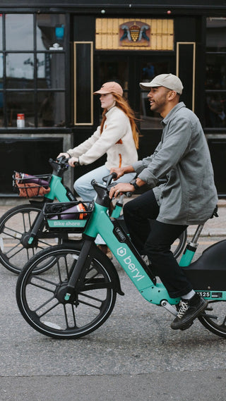 Two people riding teal Beryl bike-share bicycles on a city street, smiling and enjoying an urban ride.