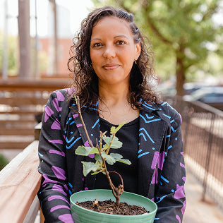 Woman holding a small potted plant outdoors, wearing a colorful patterned jacket, smiling warmly.