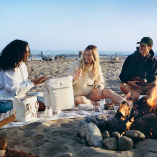 Three friends relaxing around a beach bonfire with Stanley 1913 cream cooler bag and drinkware on a picnic blanket.