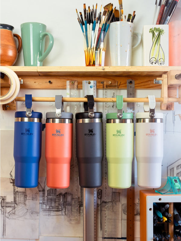 Five Stanley 1913 tumblers in blue, coral, charcoal, lime, and white hanging from a wooden shelf in an art studio.